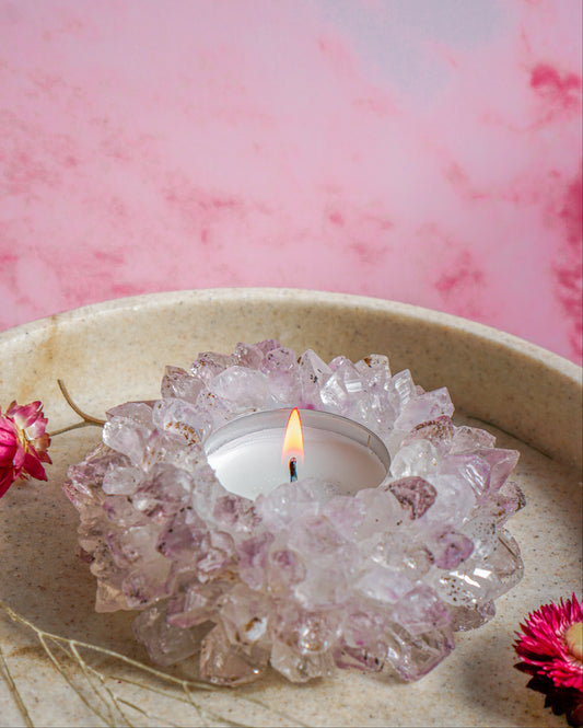 A lit tealight candle in a druzy brazilian amethyst crystal tea light holder, set in a beige ceramic dish against a pink marbled background with dried flowers.