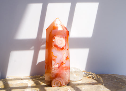 A pointed pink carnelian flower agate crystal tower made of pink peach and orange agate with white veining, standing on a stone surface next to a clear quartz crystal under direct sunlight casting shadows.