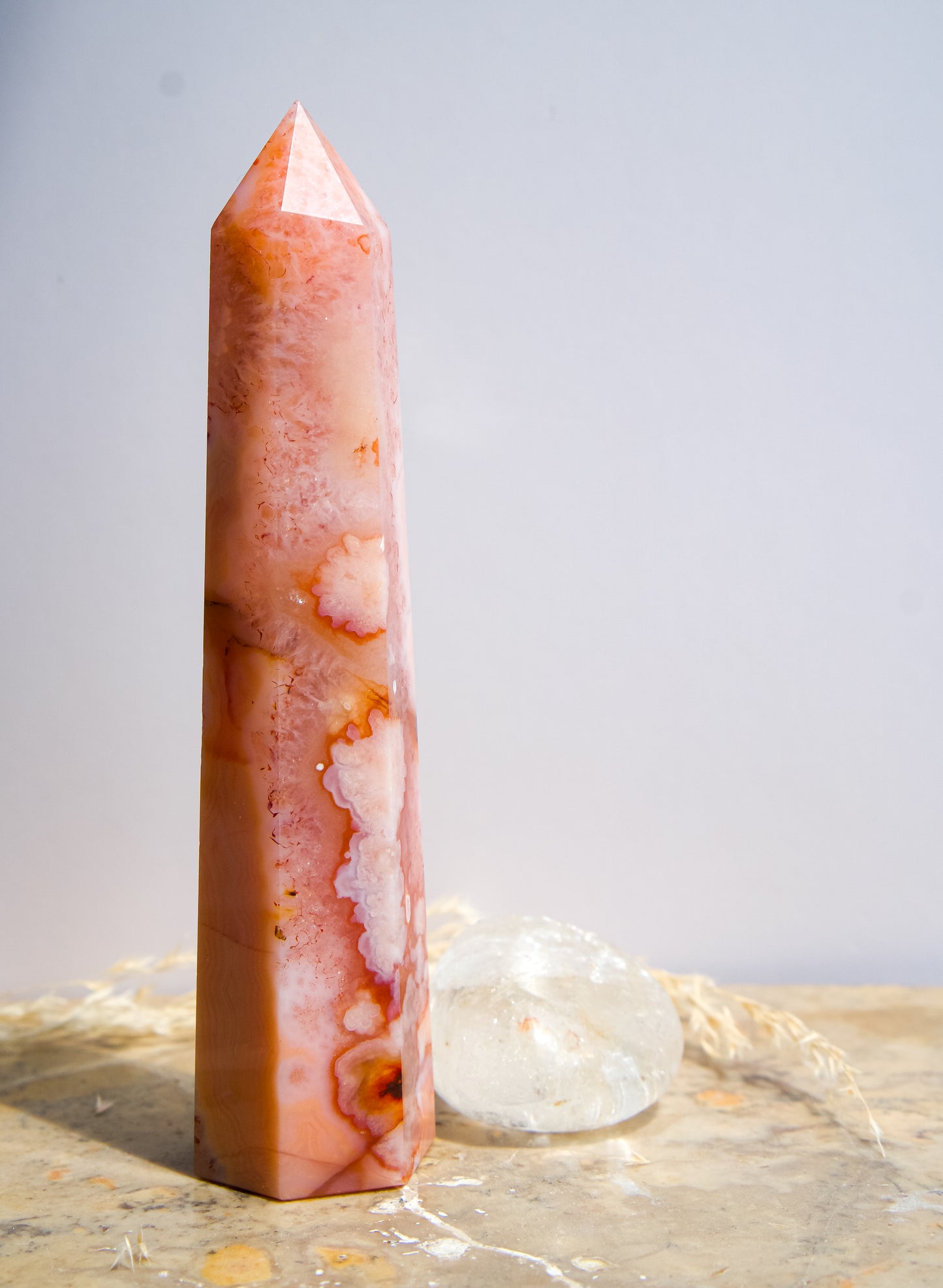 A tall, polished pink carnelian flower agate crystal tower with natural marbled patterns, standing on a textured stone surface with soft shadows cast on a white wall behind it.