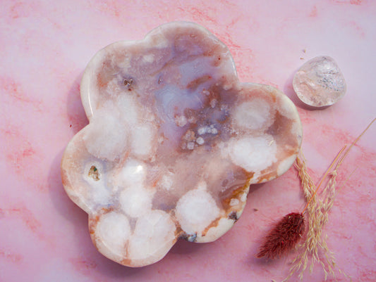 A cloud-shaped pink carnelian flower agate crystal tray with natural banded patterns in white, lavender, and brown, displayed on a pink marble surface next to a clear tumbled stone and dried grass.