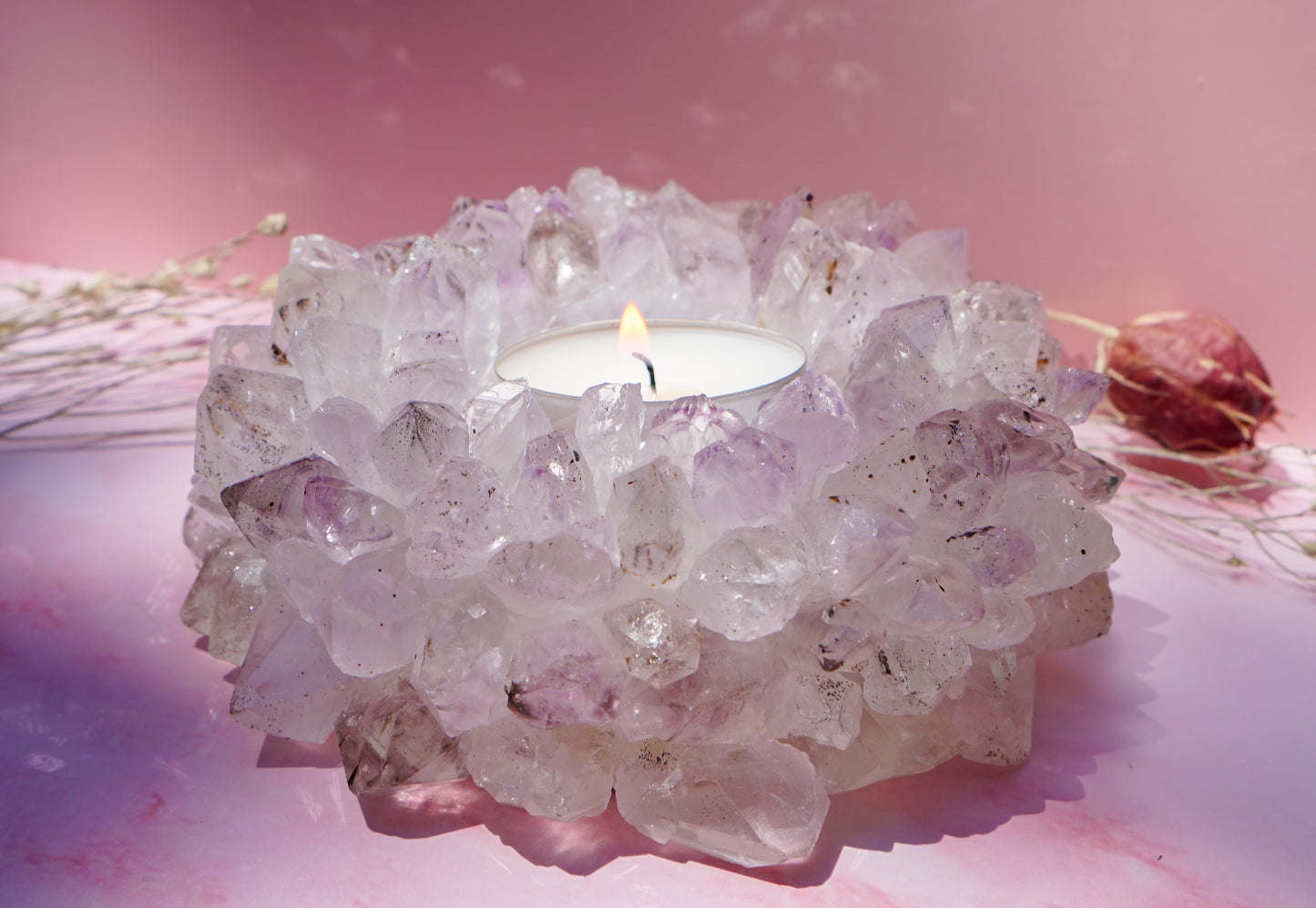A white tea light candle burning in a druzy brazilian amethyst crystal tea light holder, set against a soft pink background with dried floral props.