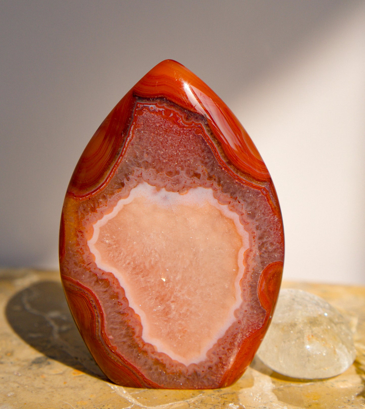 A carnelian free form flame polished teardrop-shaped agate crystal with swirling bands of red, orange, and white, standing on a textured stone surface next to a small clear quartz crystal.