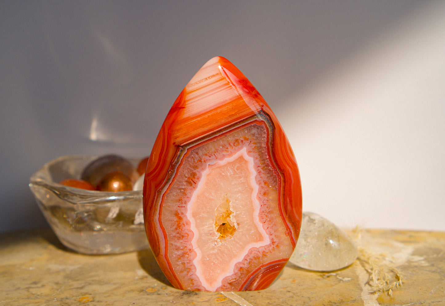 A polished freeform Carnelian flame  teardrop-shaped agate crystal with vibrant orange, red, and pink banded patterns, displayed on a stone surface next to a clear quartz crystal against a neutral gray background.
