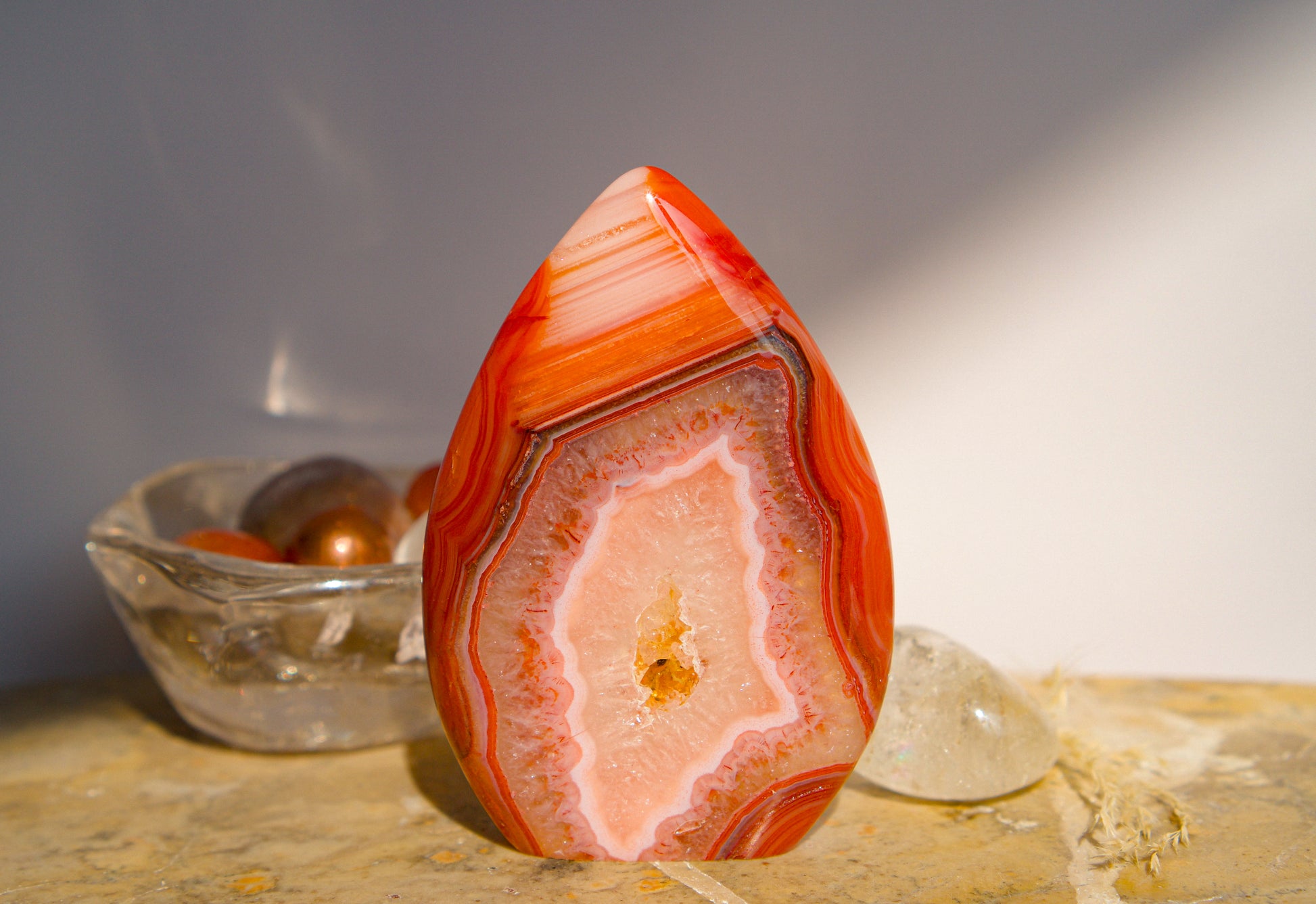 A polished freeform Carnelian flame  teardrop-shaped agate crystal with vibrant orange, red, and pink banded patterns, displayed on a stone surface next to a clear quartz crystal against a neutral gray background.