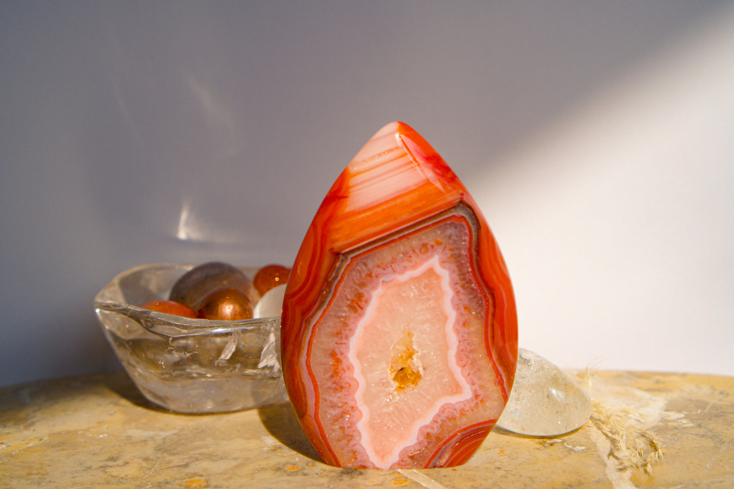 A polished freeform Carnelian flame  teardrop-shaped agate crystal with vibrant orange, red, and pink banded patterns, displayed on a stone surface next to a clear quartz crystal against a neutral gray background.