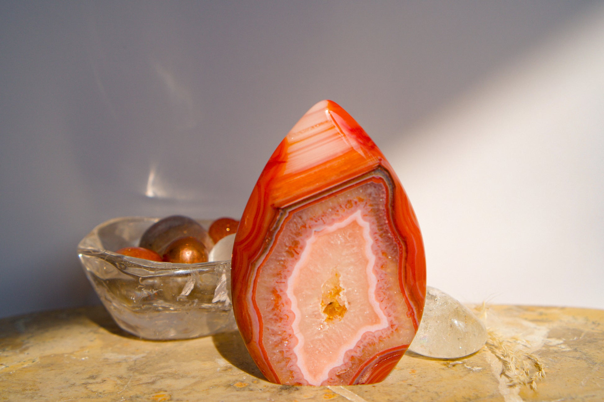 A polished freeform Carnelian flame  teardrop-shaped agate crystal with vibrant orange, red, and pink banded patterns, displayed on a stone surface next to a clear quartz crystal against a neutral gray background.