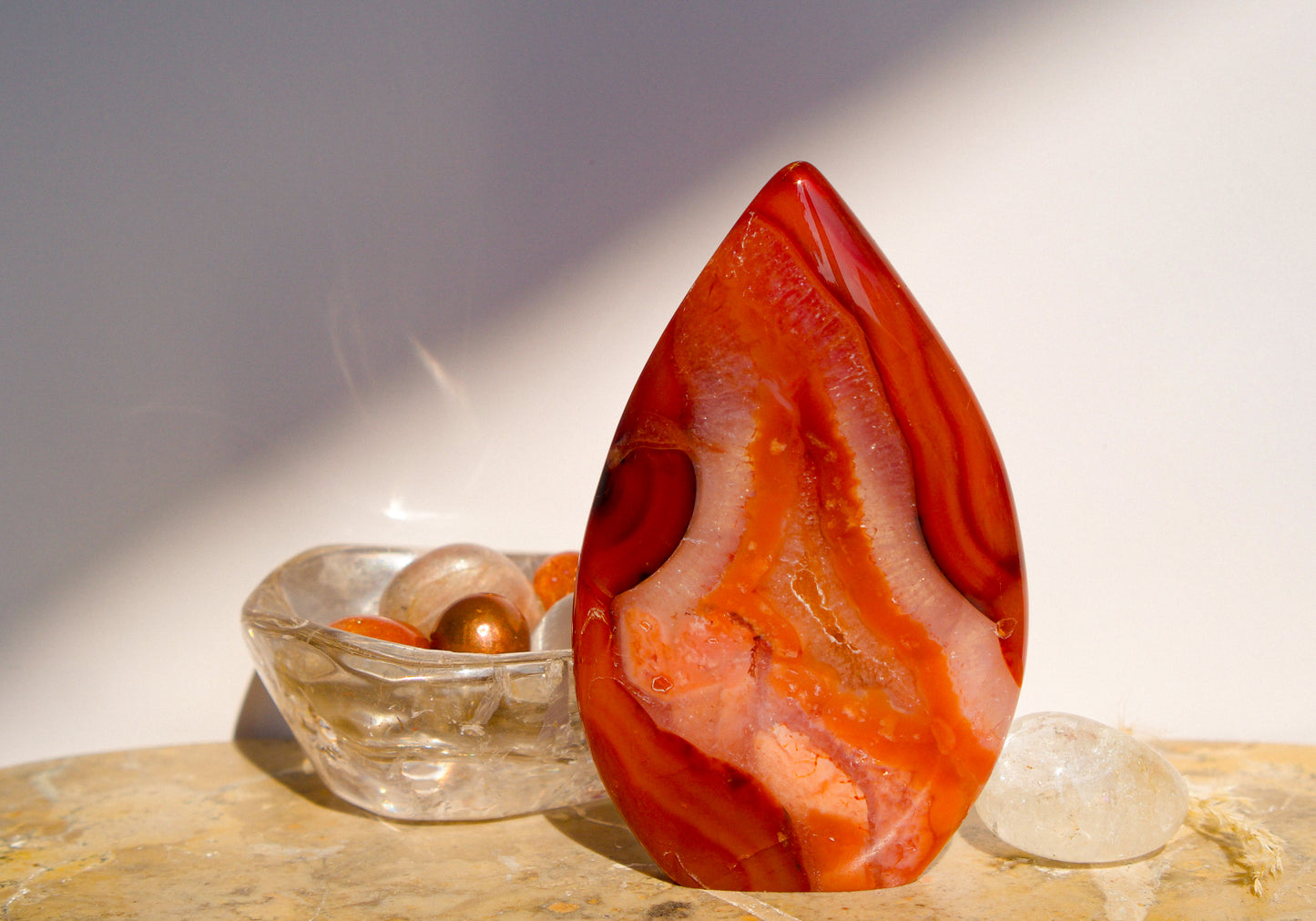 A polished freeform Carnelian flame  teardrop-shaped agate crystal with vibrant orange, red, and pink banded patterns, displayed on a stone surface next to a clear quartz crystal against a neutral gray background.