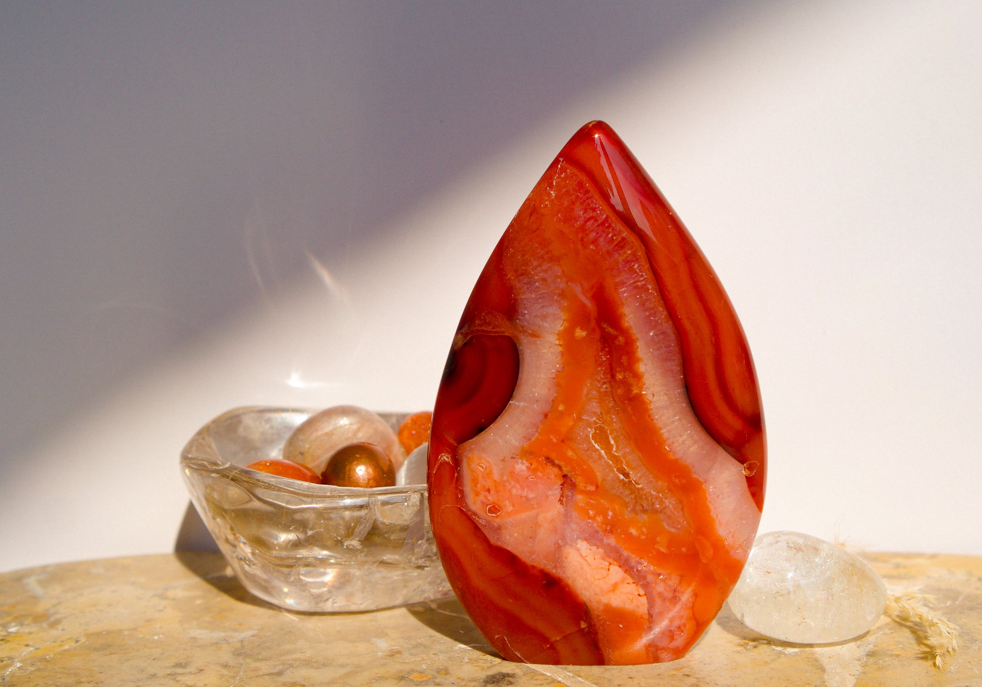 A polished freeform Carnelian flame  teardrop-shaped agate crystal with vibrant orange, red, and pink banded patterns, displayed on a stone surface next to a clear quartz crystal against a neutral gray background.