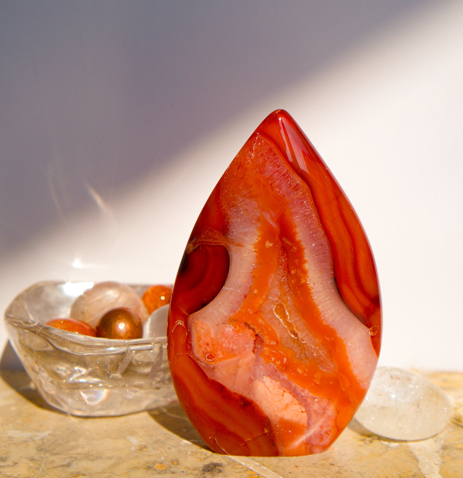 A polished freeform Carnelian flame  teardrop-shaped agate crystal with vibrant orange, red, and pink banded patterns, displayed on a stone surface next to a clear quartz crystal against a neutral gray background.