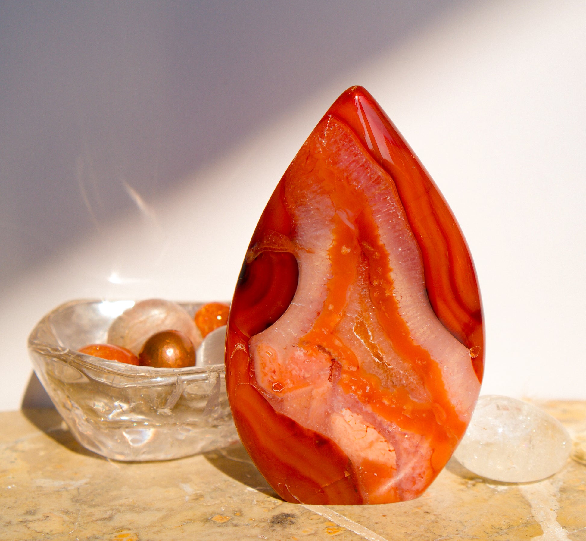 A polished freeform Carnelian flame  teardrop-shaped agate crystal with vibrant orange, red, and pink banded patterns, displayed on a stone surface next to a clear quartz crystal against a neutral gray background.