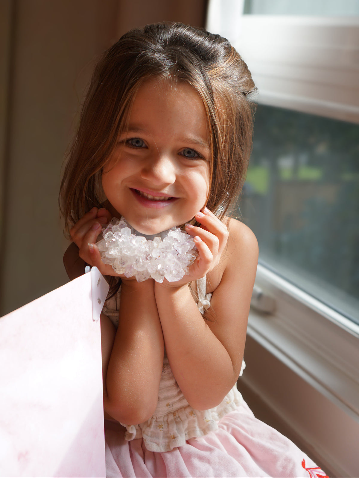 A young girl with brown hair smiles while holding a Brazilian amethyst teal ight near her face, wearing a light pink dress in front of a window.