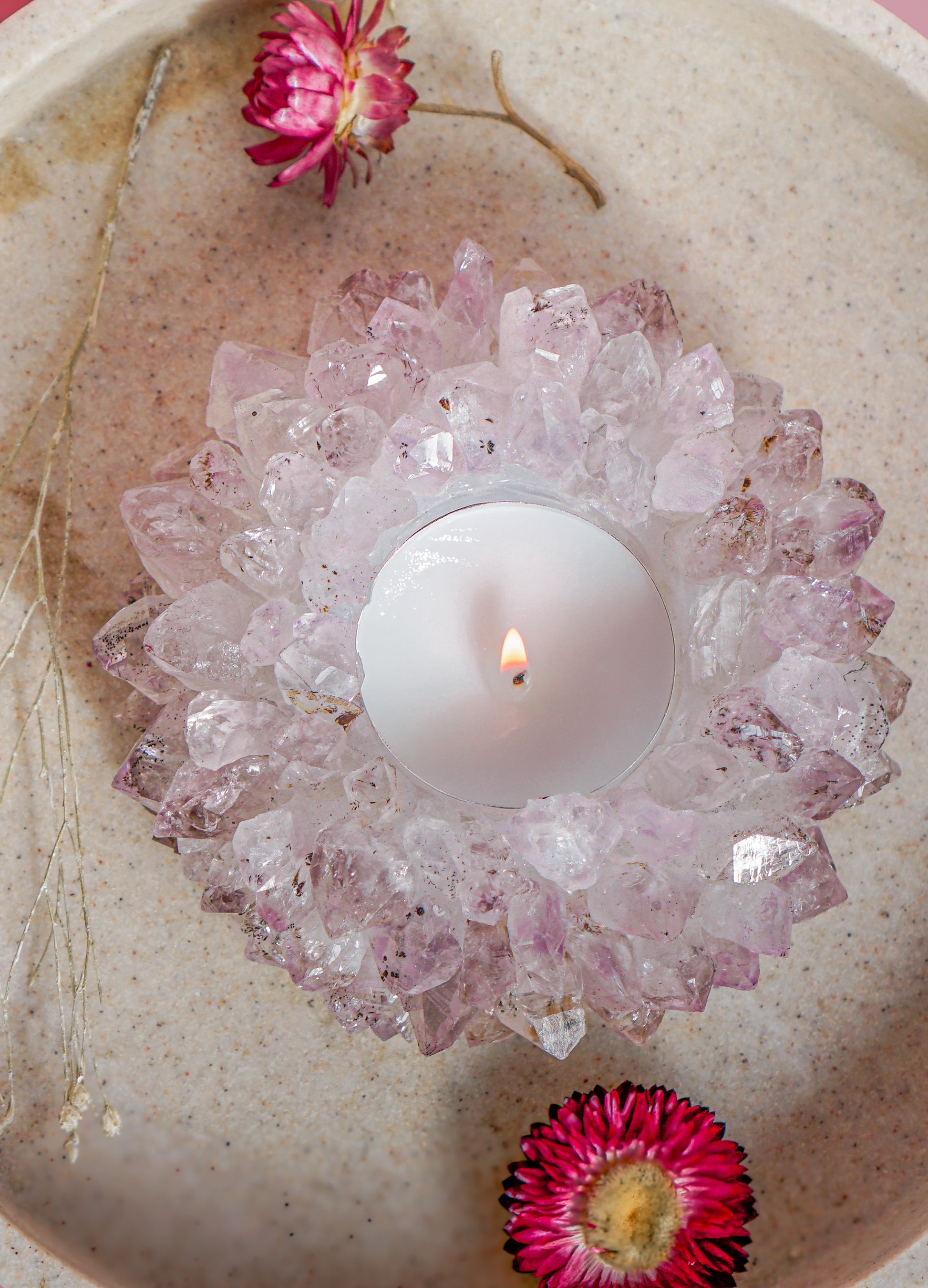 A lit white tea light candle in a druzy brazilian amethyst crystal tea light holder, placed in a beige speckled bowl with dried flowers.