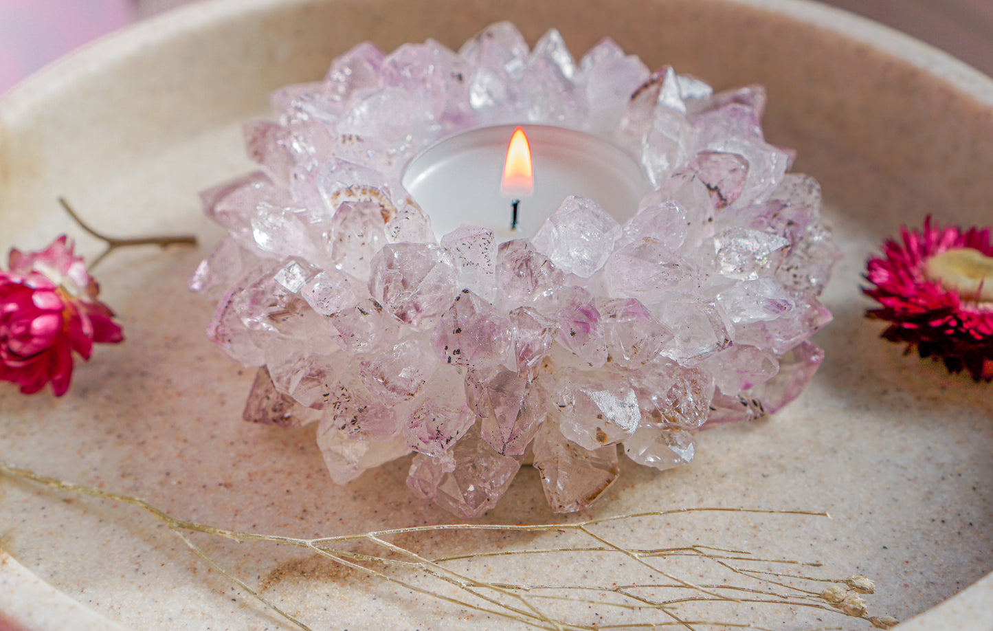 A lit tealight candle in a druzy brazilian amethyst crystal tea light holder styled on a beige surface with small pink flowers around it.