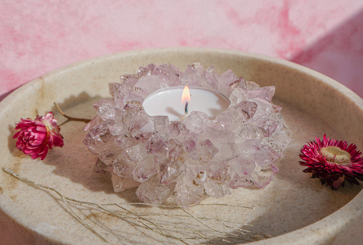 A lit white tea light candle set idruzy brazilian amethyst crystal tea light holder, placed in a beige stone dish with dried pink flowers on a pink background.