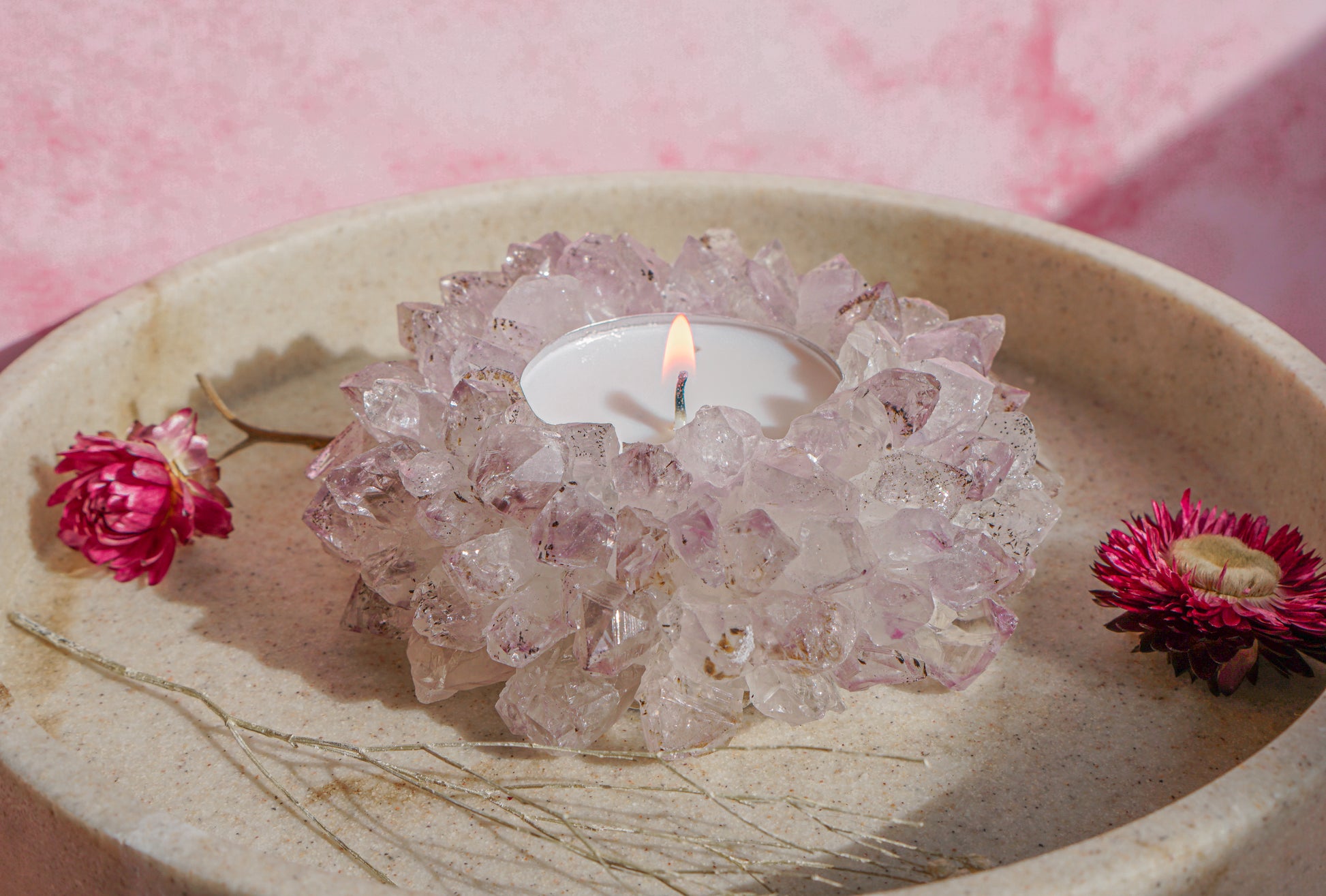 A lit white tea light candle set idruzy brazilian amethyst crystal tea light holder, placed in a beige stone dish with dried pink flowers on a pink background.