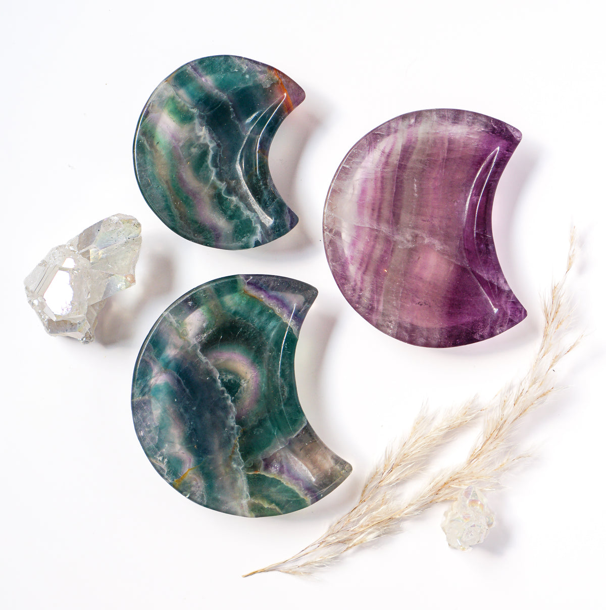 Three carved fluorite crescent moon dishes in teal-violet, amber-green, and lavender purple tones, styled with a raw quartz cluster and dried pampas grass