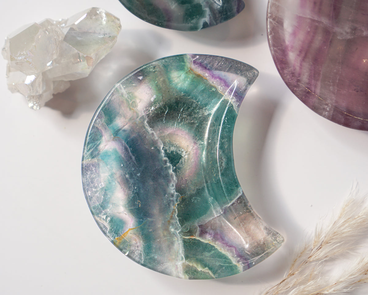 Rainbow fluorite crescent moon dish with a teal and violet circular banded eye pattern, styled beside a quartz crystal cluster, another fluorite moon dish, and dried pampas grass
