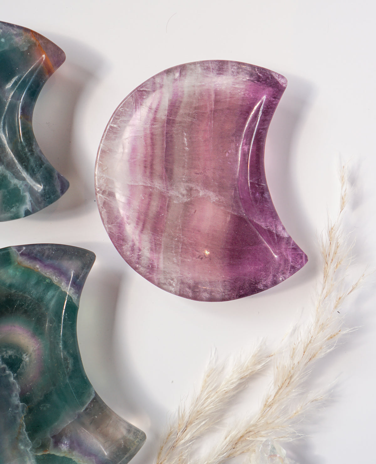 Purple fluorite crescent moon dish beside two rainbow fluorite crescent moon dishes in teal, green, and amber tones, with dried pampas grass