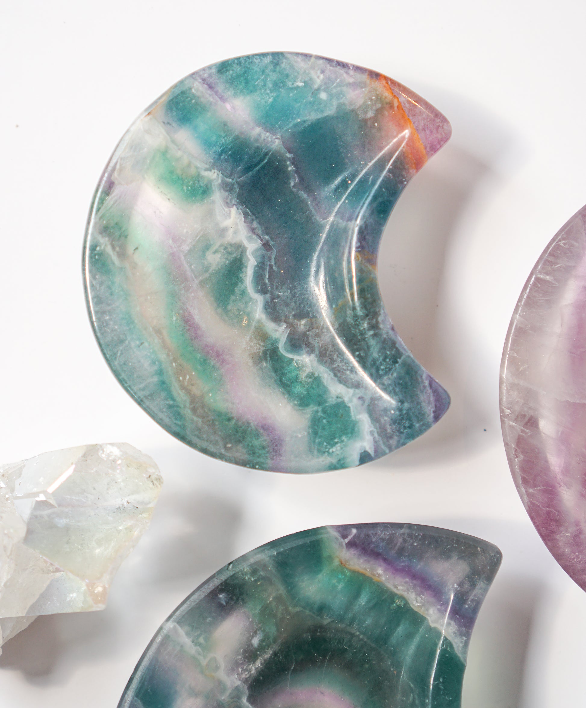 Rainbow fluorite crescent moon dish with teal-green and amber banding, displayed beside a teal-violet fluorite moon dish, a purple fluorite moon dish, and a raw quartz cluster