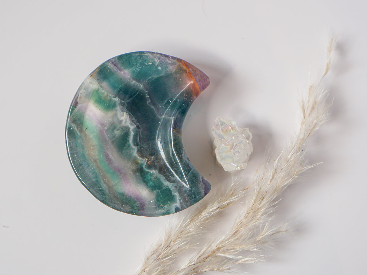 Rainbow fluorite crescent moon dish in teal-green with amber banding, styled with a raw quartz cluster and dried pampas grass