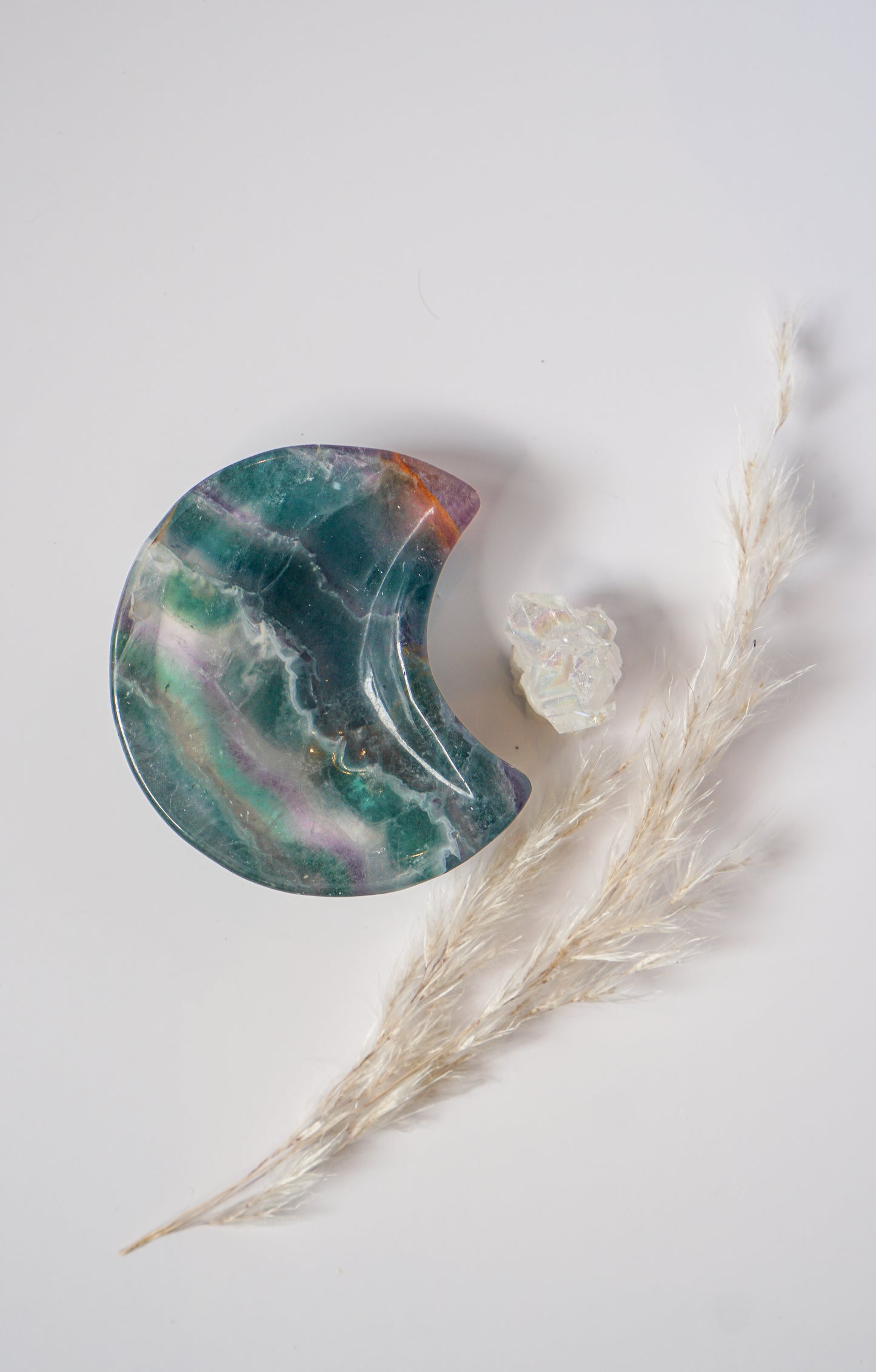 Vertical view of a rainbow fluorite crescent moon dish in teal-green with amber banding, beside a raw quartz cluster and dried pampas grass
