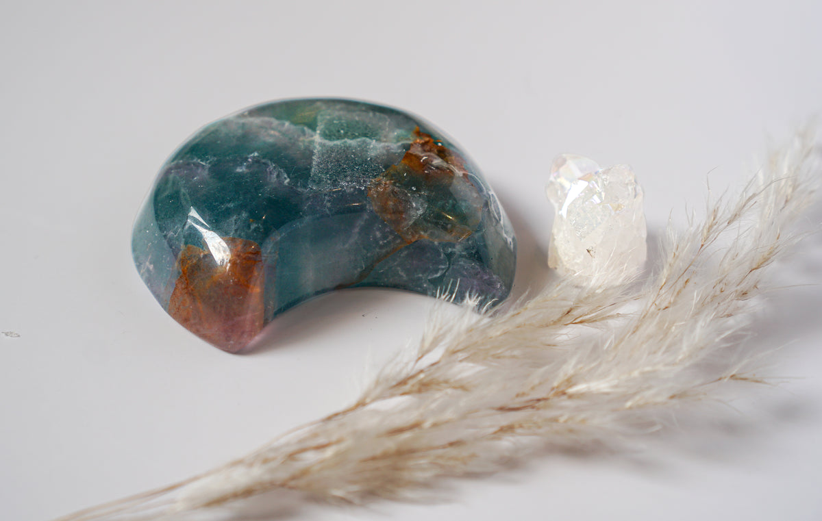 Close-up of a rainbow fluorite crescent moon dish showing teal-green stone with rich amber and copper banding, beside a raw quartz cluster and dried pampas grass