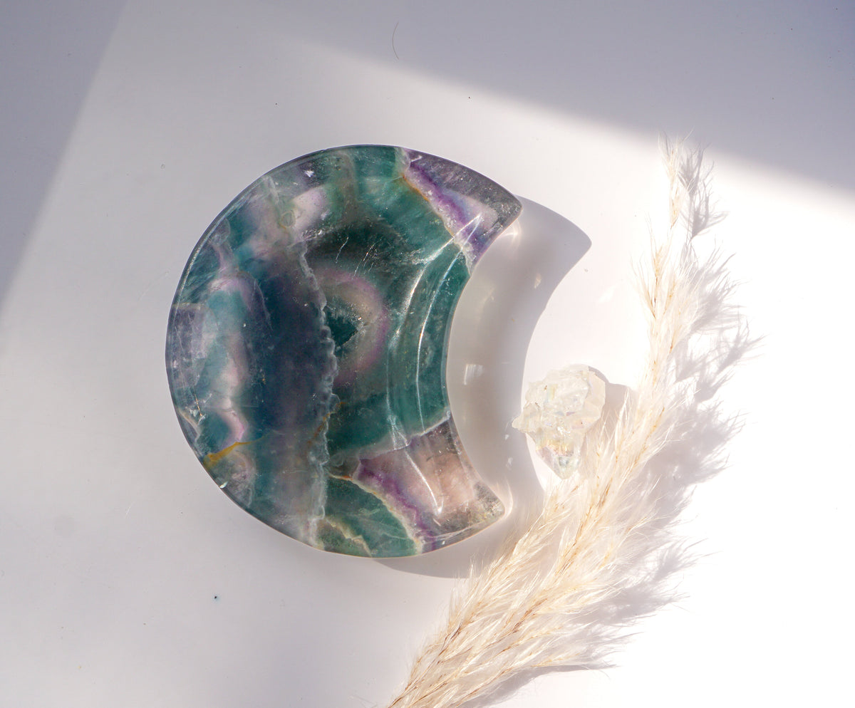 Close-up of a rainbow fluorite crescent moon dish in teal and violet with a circular banded eye pattern, in natural sunlight beside a raw quartz point and dried pampas grass