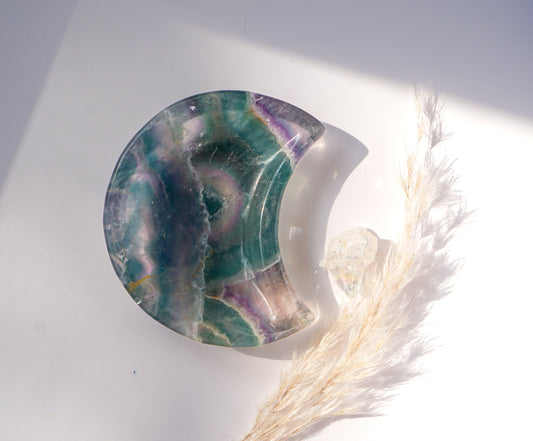 Close-up of a rainbow fluorite crescent moon dish in teal and violet with a circular banded eye pattern, in natural sunlight beside a raw quartz point and dried pampas grass