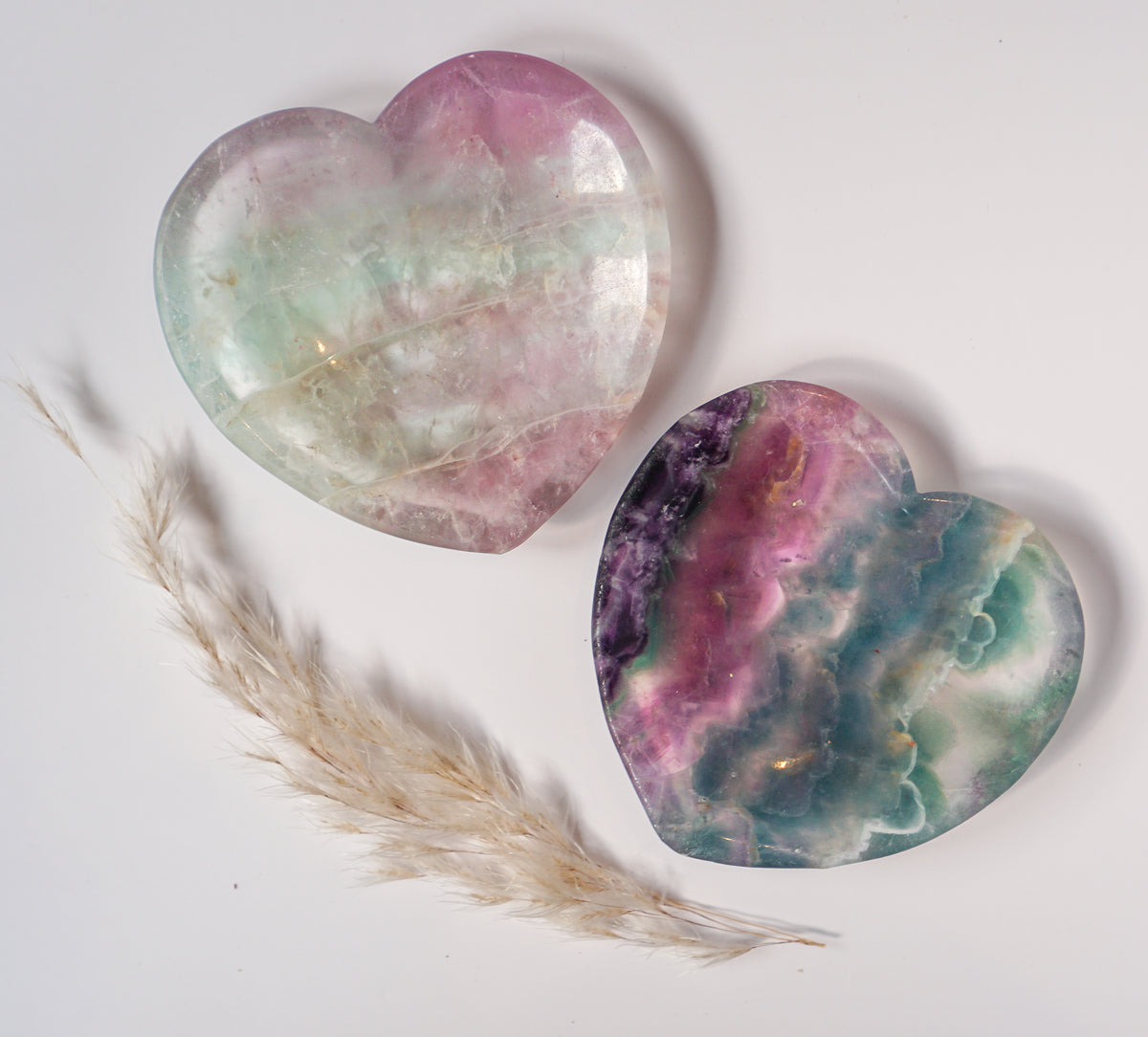 Two carved rainbow fluorite heart dishes together, one in soft mint and pink and one in bold purple, pink, and teal, with dried pampas grass