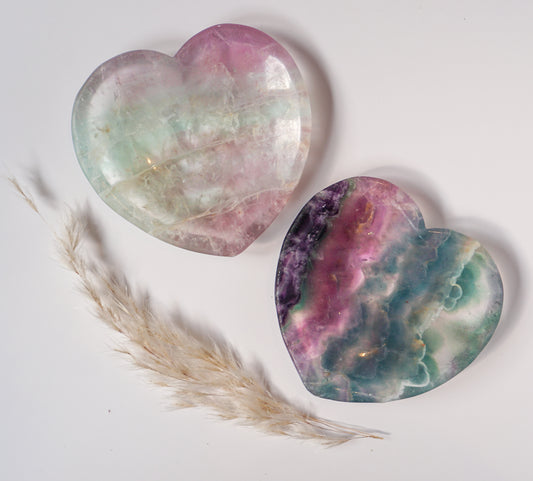 Two carved rainbow fluorite heart dishes together, one in soft mint and pink and one in bold purple, pink, and teal, with dried pampas grass