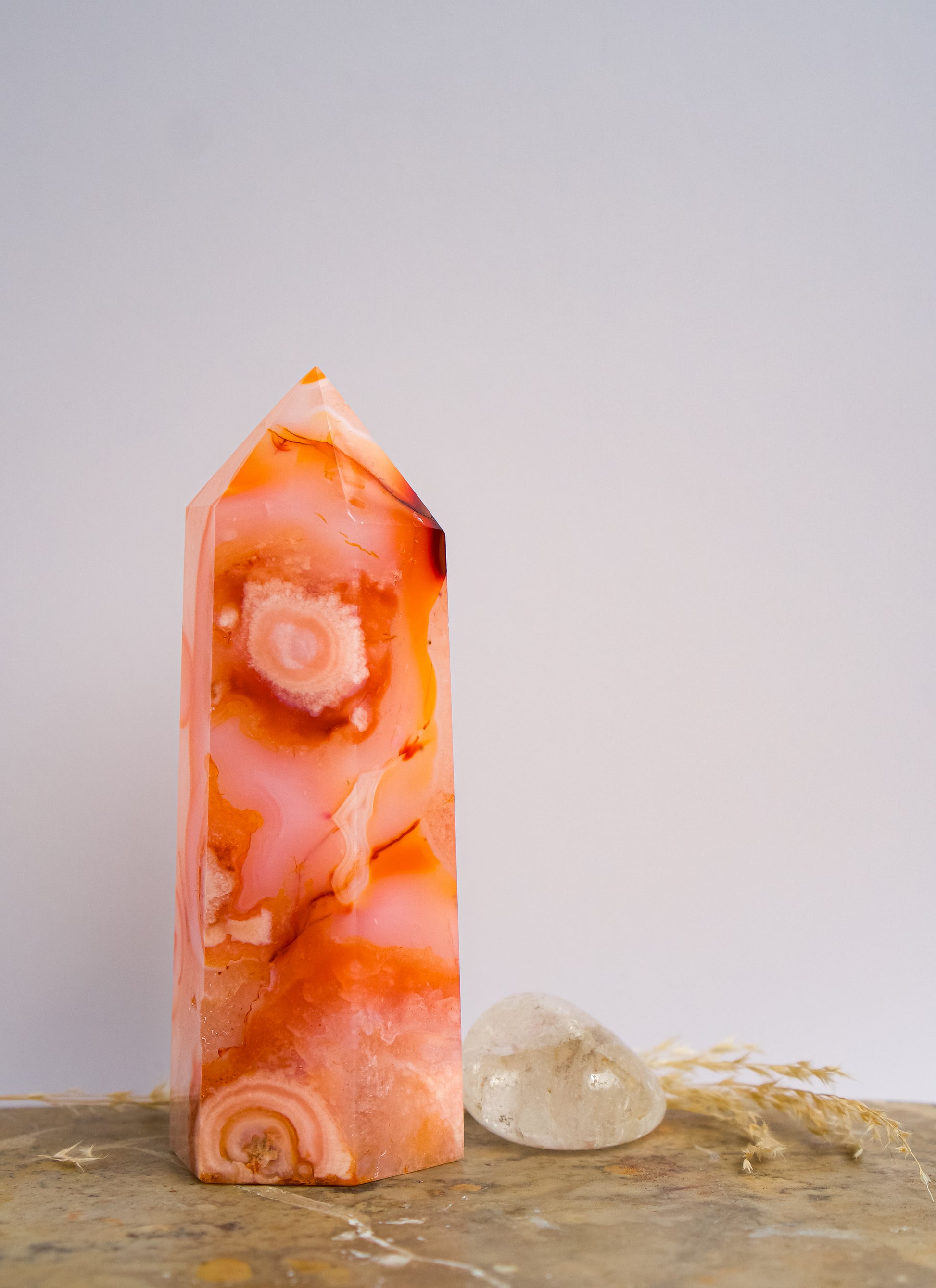 A pointed pink carnelian flower agate crystal tower made of  pink peach and orange agate with white veining, standing on a stone surface next to a clear quartz crystal under direct sunlight casting shadows.