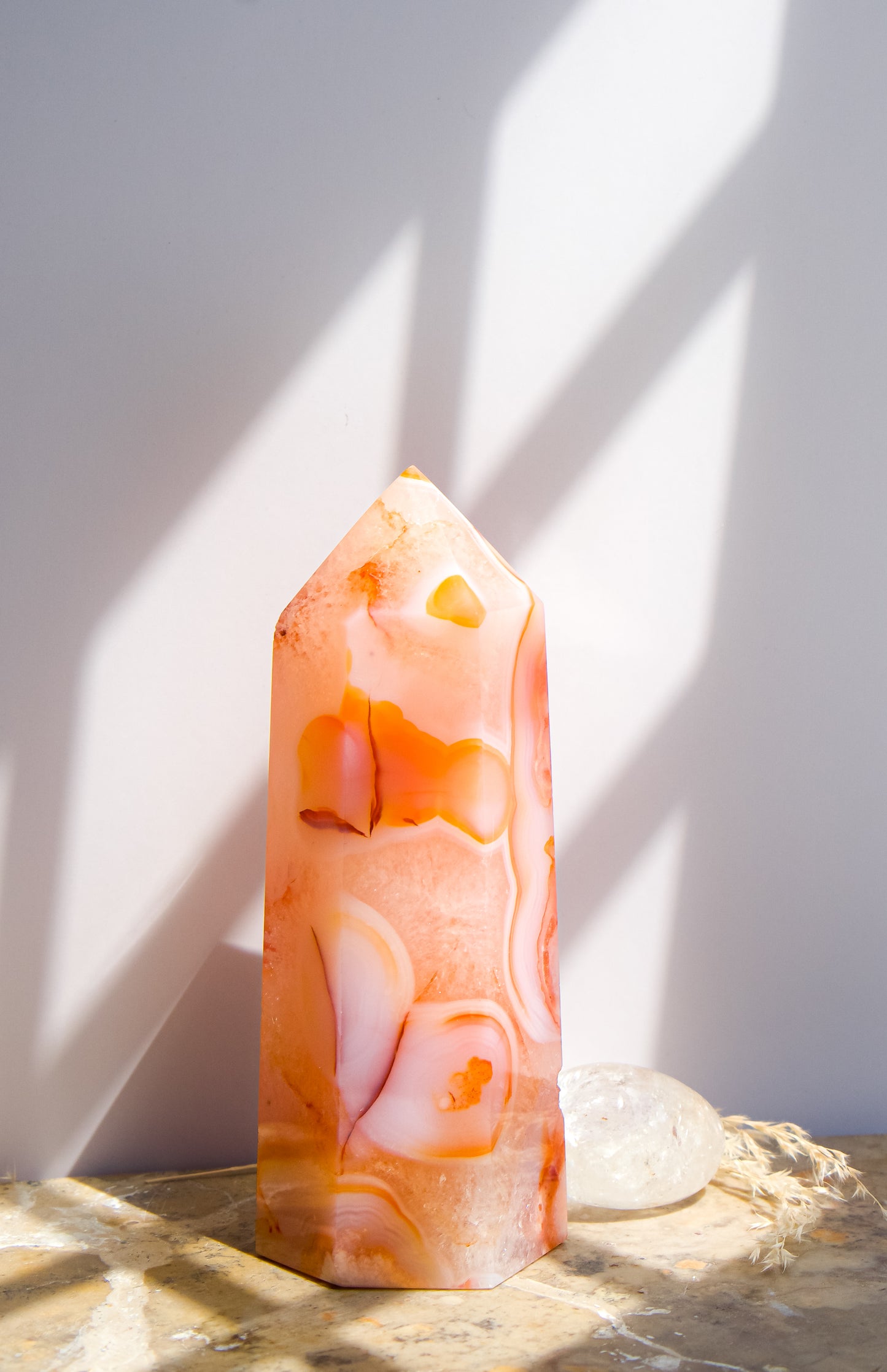 A pointed pink carnelian flower agate crystal tower made of  pink peach and orange agate with white veining, standing on a stone surface next to a clear quartz crystal under direct sunlight casting shadows.