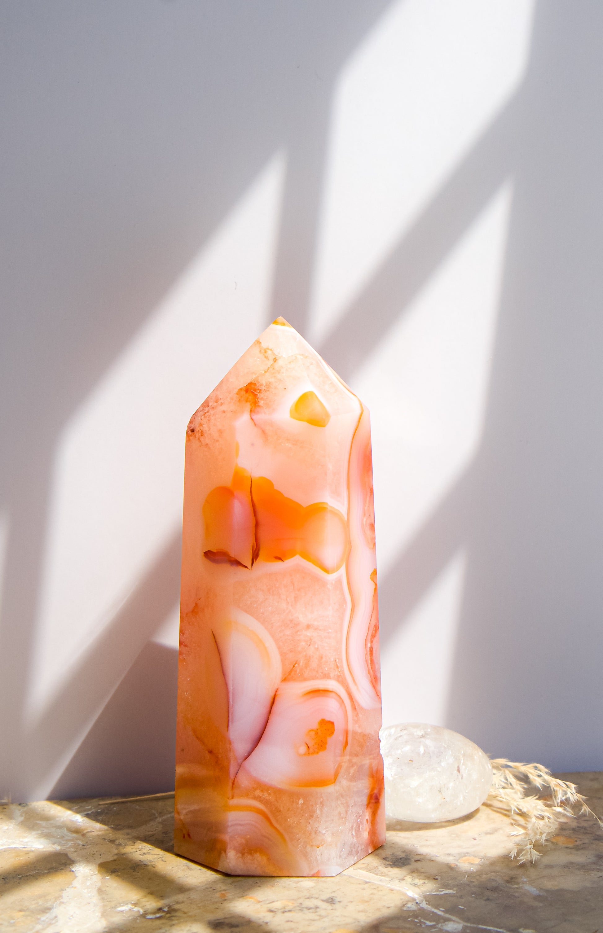 A pointed pink carnelian flower agate crystal tower made of  pink peach and orange agate with white veining, standing on a stone surface next to a clear quartz crystal under direct sunlight casting shadows.