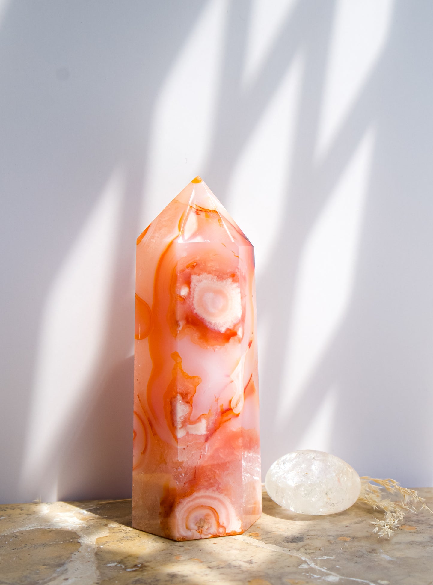 A pointed pink carnelian flower agate crystal tower made of  pink peach and orange agate with white veining, standing on a stone surface next to a clear quartz crystal under direct sunlight casting shadows.