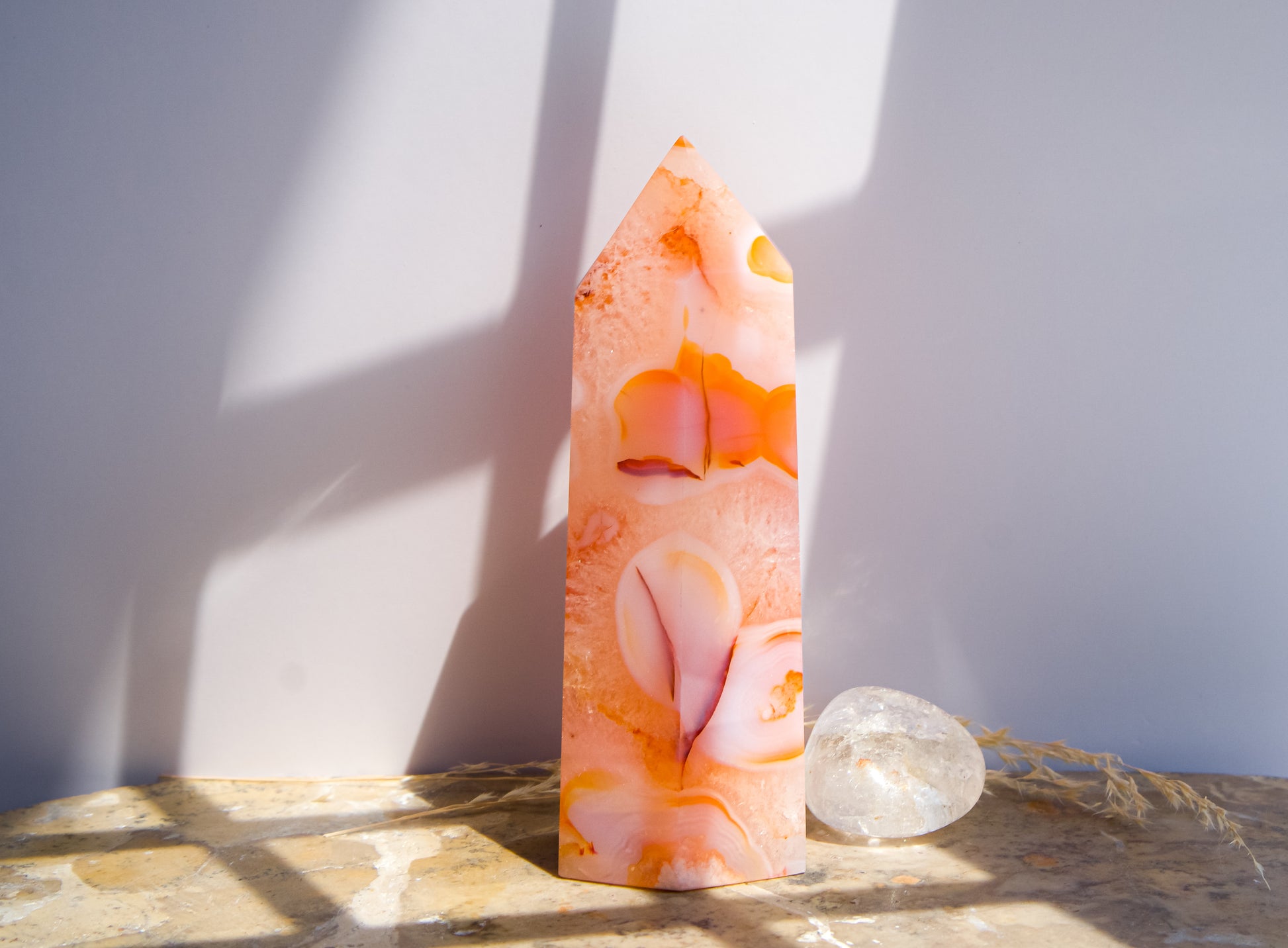 A pointed pink carnelian flower agate crystal tower made of  pink peach and orange agate with white veining, standing on a stone surface next to a clear quartz crystal under direct sunlight casting shadows.