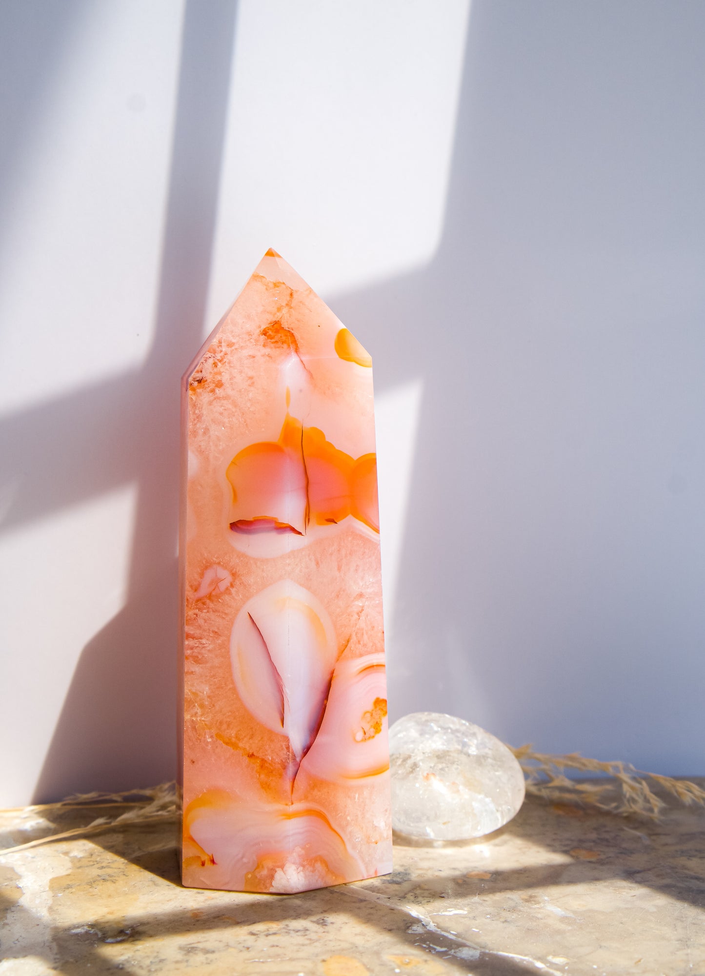 A pointed pink carnelian flower agate crystal tower made of  pink peach and orange agate with white veining, standing on a stone surface next to a clear quartz crystal under direct sunlight casting shadows.