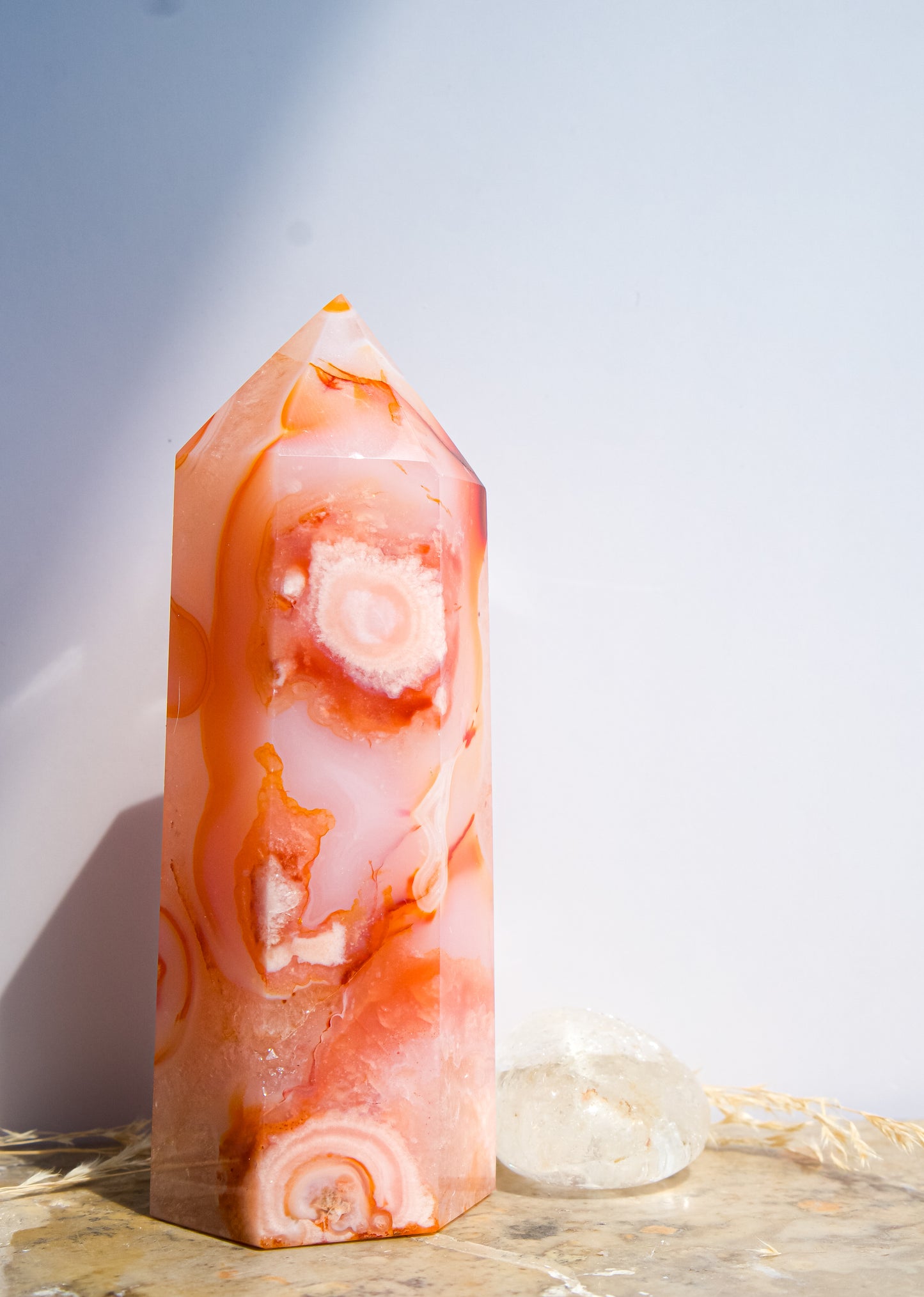 A pointed pink carnelian flower agate crystal tower made of  pink peach and orange agate with white veining, standing on a stone surface next to a clear quartz crystal under direct sunlight casting shadows.
