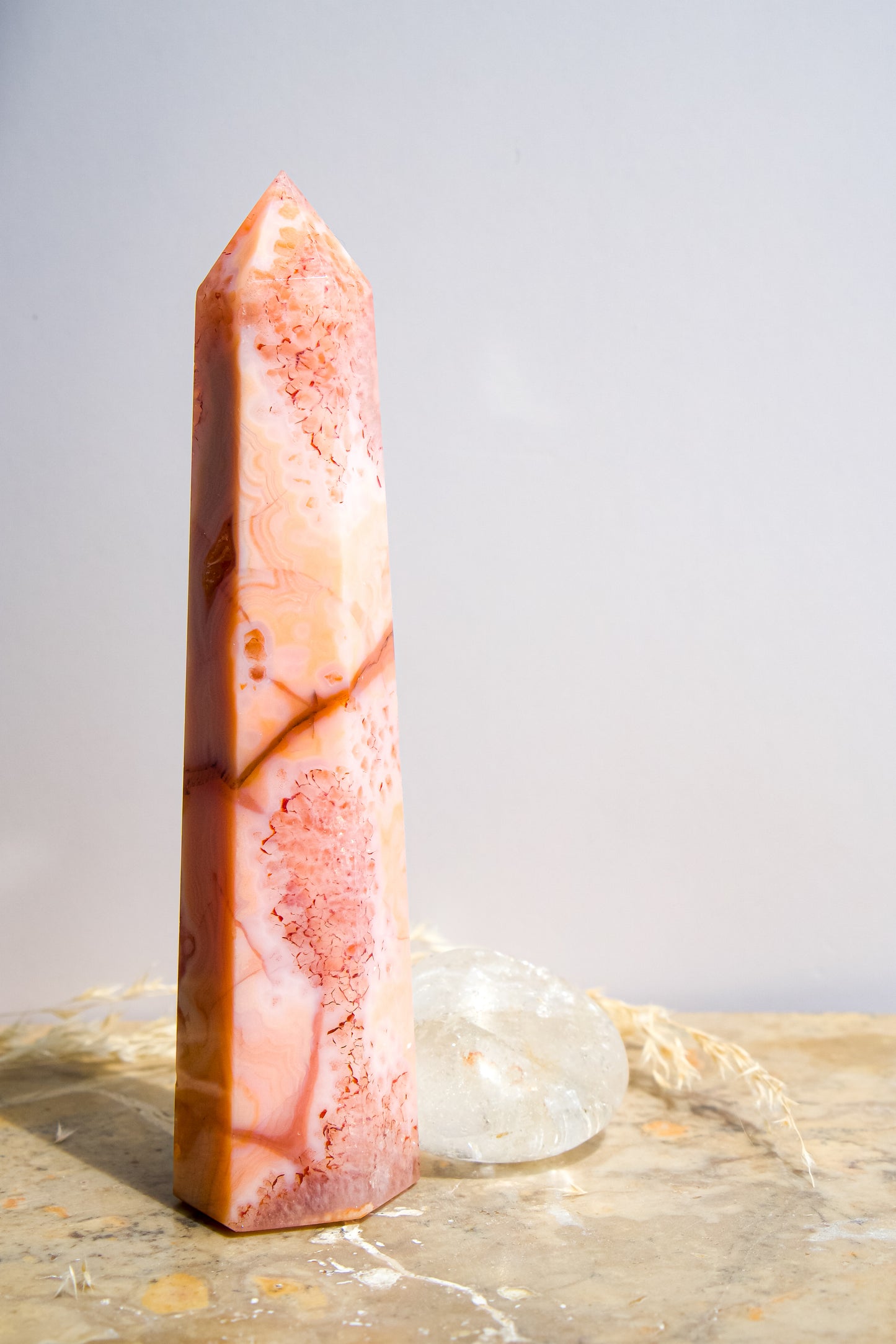 A tall, polished pink carnelian flower agate crystal tower with natural marbled patterns, standing on a textured stone surface with soft shadows cast on a white wall behind it.
