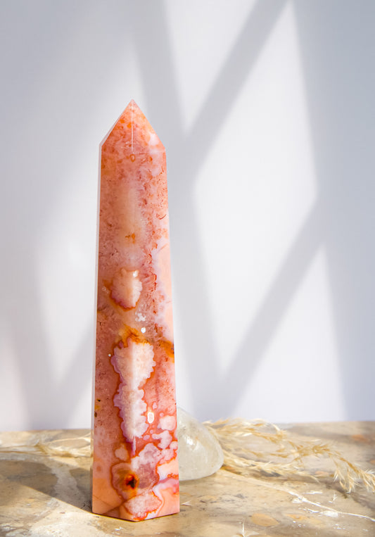 A tall, polished pink carnelian flower agate crystal tower with natural marbled patterns, standing on a textured stone surface with soft shadows cast on a white wall behind it.