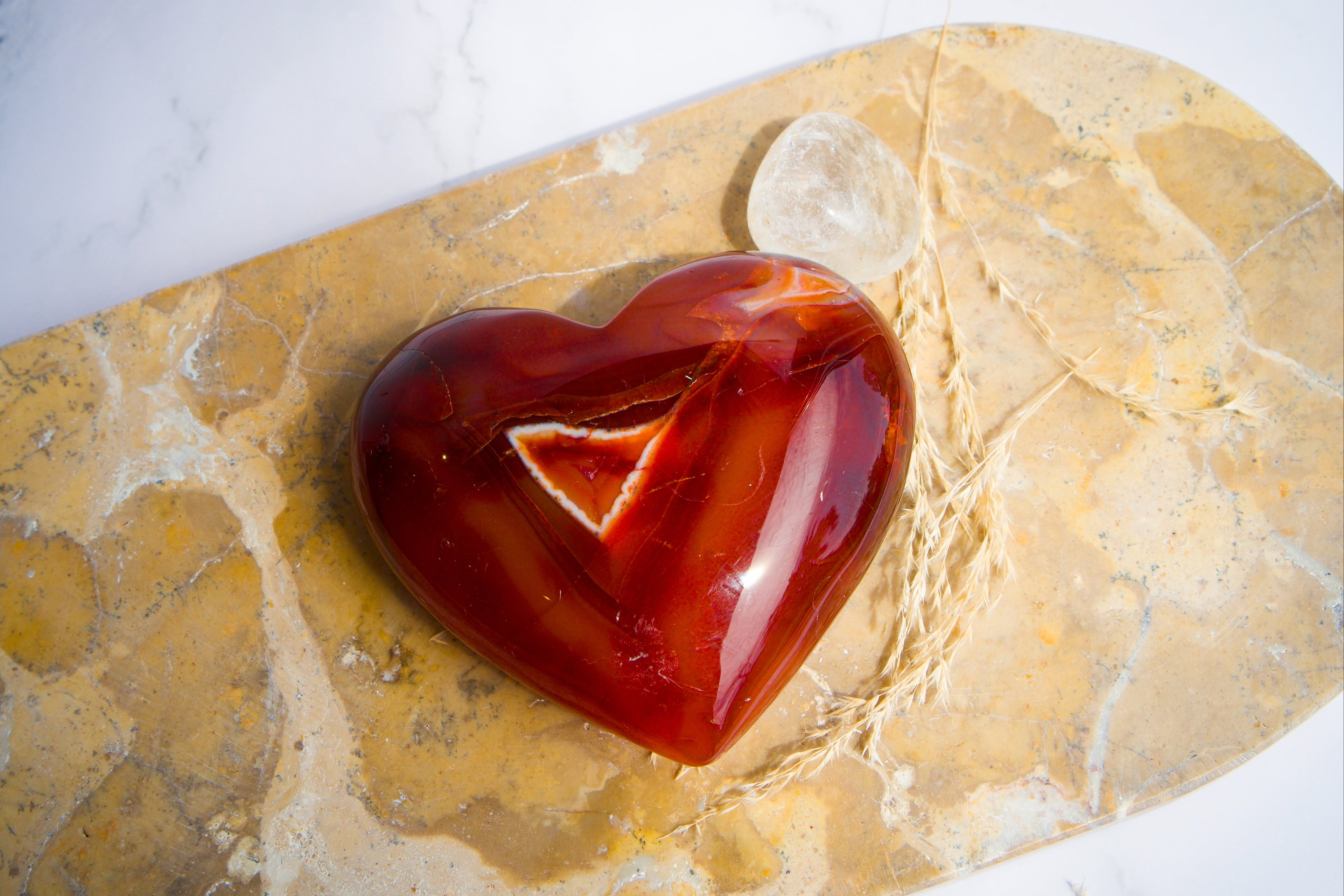 A polished red carnelian heart-shaped agate stone ornament with a triangular cutout, displayed on a yellowish stone slab with a small clear crystal beside it.