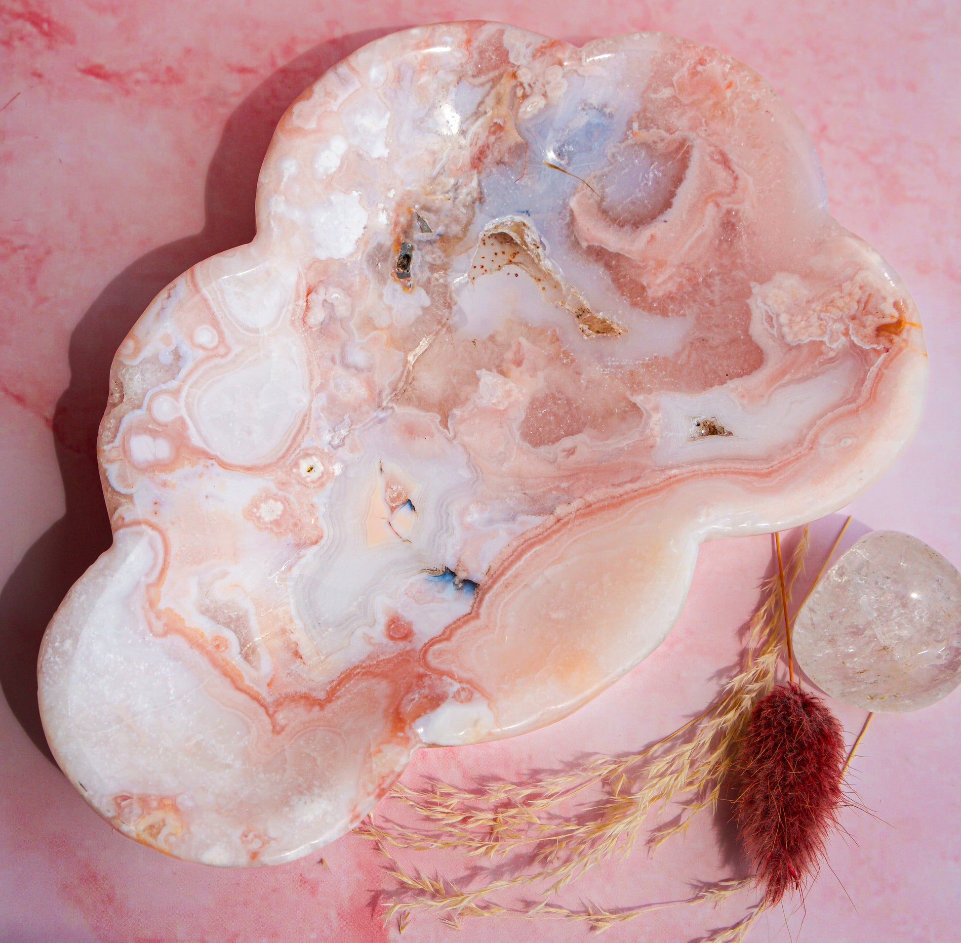 A polished pink carnelian flower agate stone tray with organic cloud-like shapes and pink, white, and peach banding, styled on a pink surface with dried grass and a clear quartz crystal.