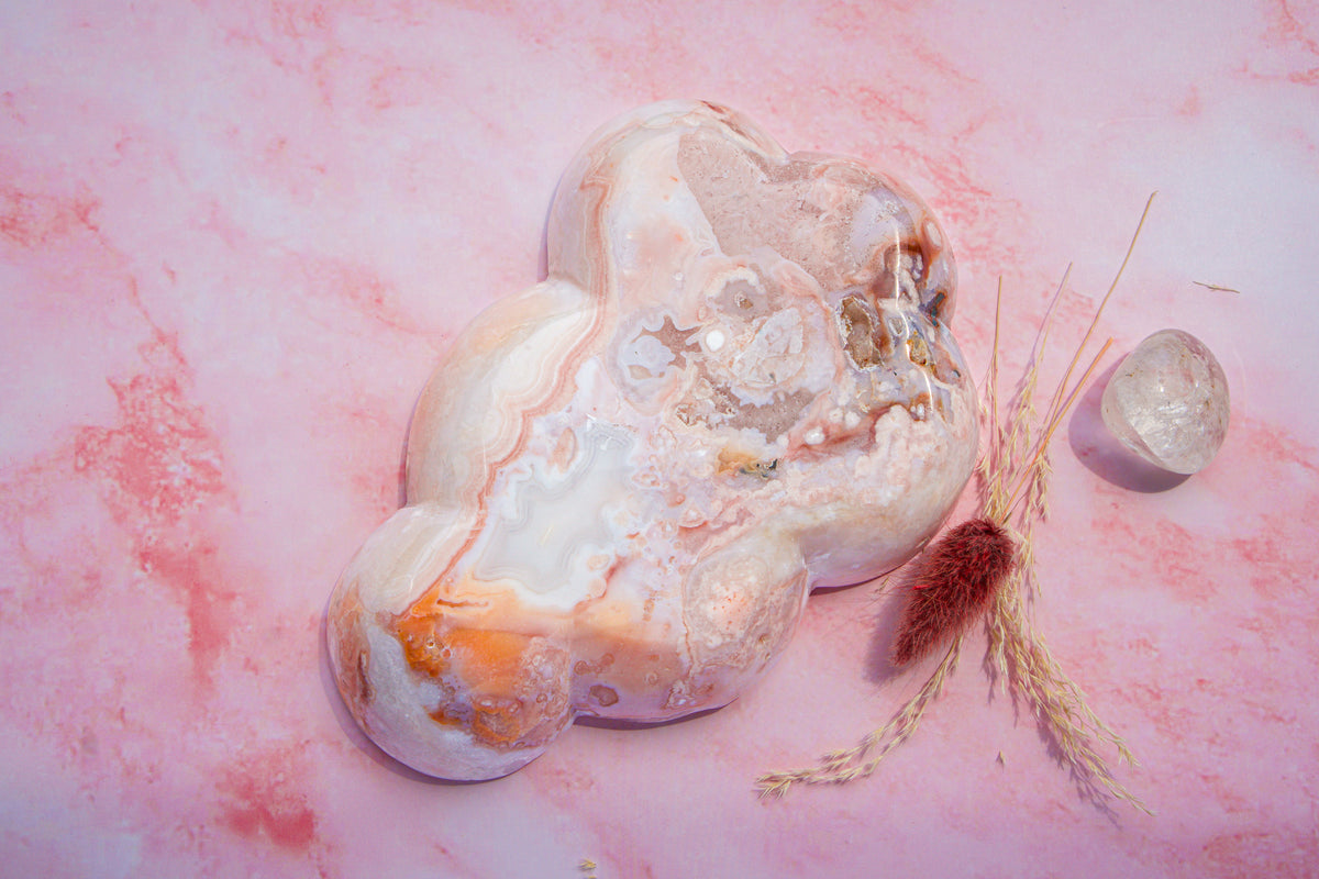 A polished irregular-shaped pink carnelian flower agate stone with swirling bands of white, peach, and orange, displayed on a pink marbled surface next to a small clear crystal and dried grass.