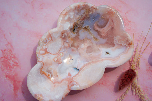 A cloud-shaped dish made from polished carnerlian flower agate stone with natural pink, white, and beige banded patterns, displayed on a pink marble surface next to dried grass.