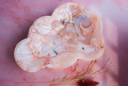 A polished cloud-shaped  pink carnlian flower agate stone with pink, white, and pale blue marbled patterns, displayed on a pink marbled surface with dried grass.