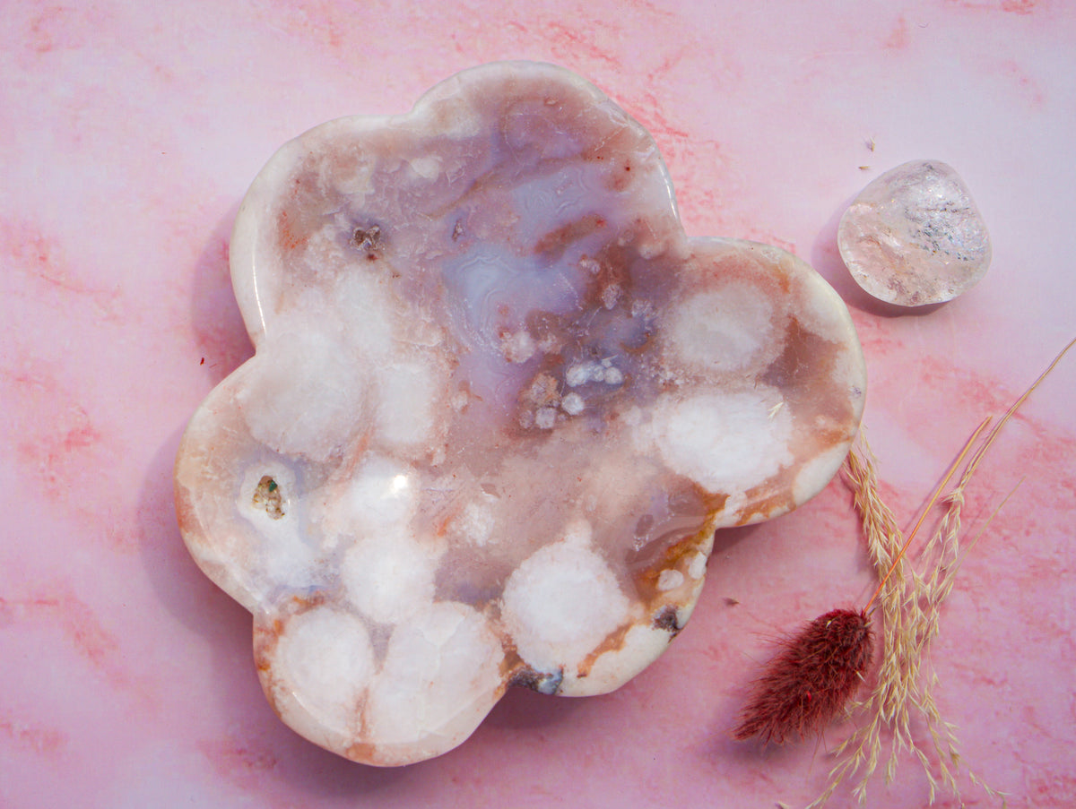 A cloud-shaped  pink carnelian flower agate crystal tray with natural banded patterns in white, lavender, and brown, displayed on a pink marble surface next to a clear tumbled stone and dried grass.