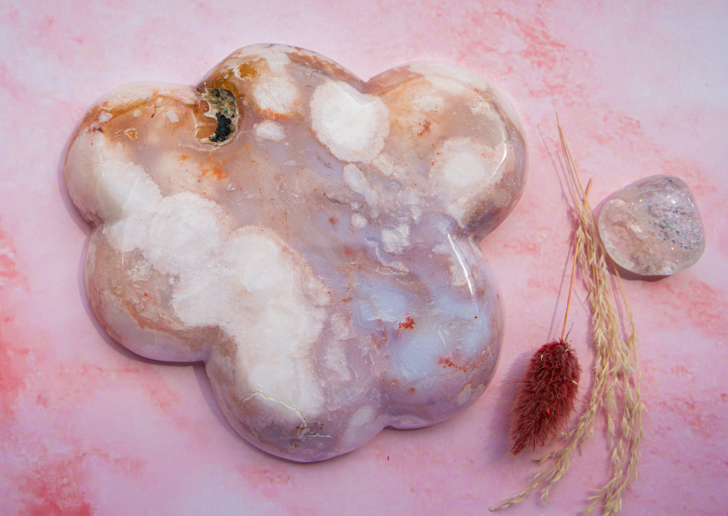A polished, cloud-shaped  pink carnelian flower agate crystal tray with marbled white, lavender, and peach tones, displayed on a pink marbled surface next to dried grass and a smaller clear crystal.