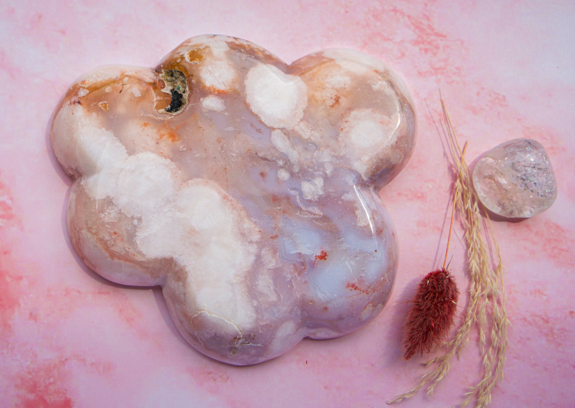 A polished, cloud-shaped  pink carnelian flower agate crystal tray with marbled white, lavender, and peach tones, displayed on a pink marbled surface next to dried grass and a smaller clear crystal.