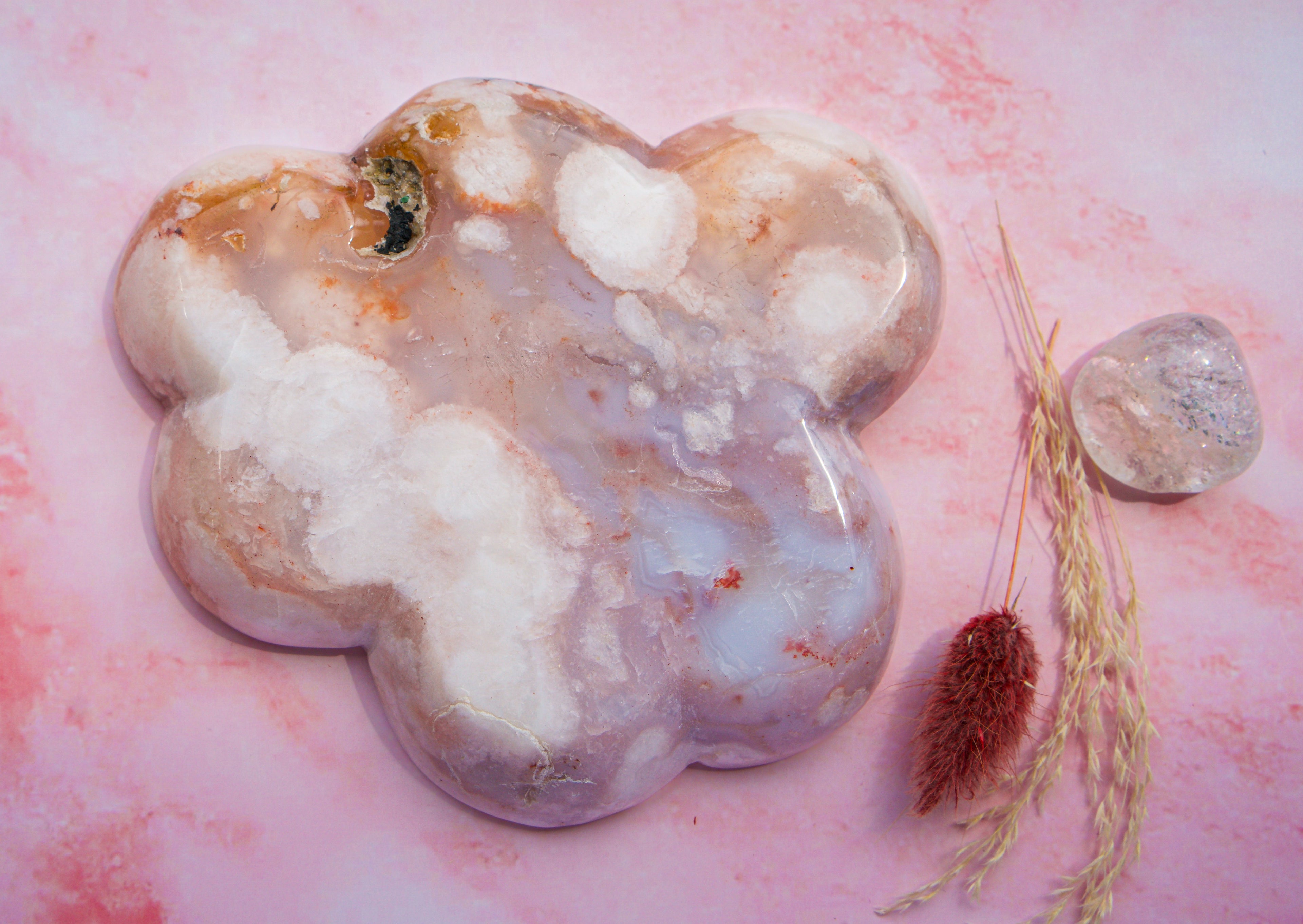 A polished, cloud-shaped  pink carnelian flower agate crystal tray with marbled white, lavender, and peach tones, displayed on a pink marbled surface next to dried grass and a smaller clear crystal.