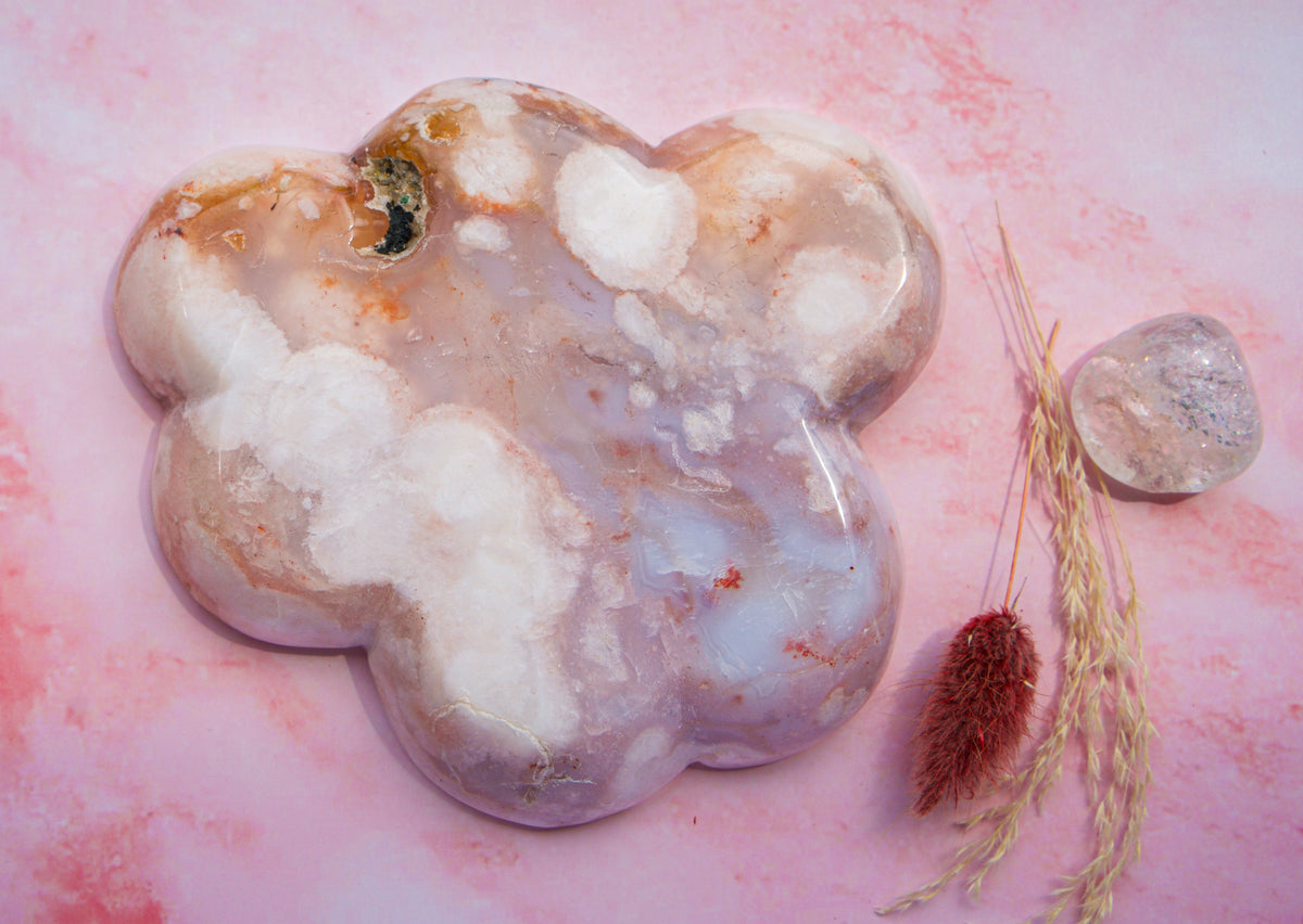 A polished, cloud-shaped  pink carnelian flower agate crystal stone with marbled white, lavender, and peach tones, displayed on a pink marbled surface next to dried grass and a smaller clear crystal.