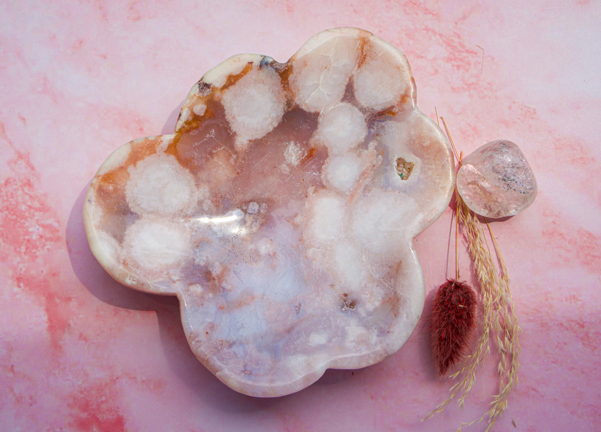 A cloud shaped  pink carnelian flower agate tray white and orange crystalline patterns, displayed on a pink marbled surface next to a small clear crystal and dried grass.
